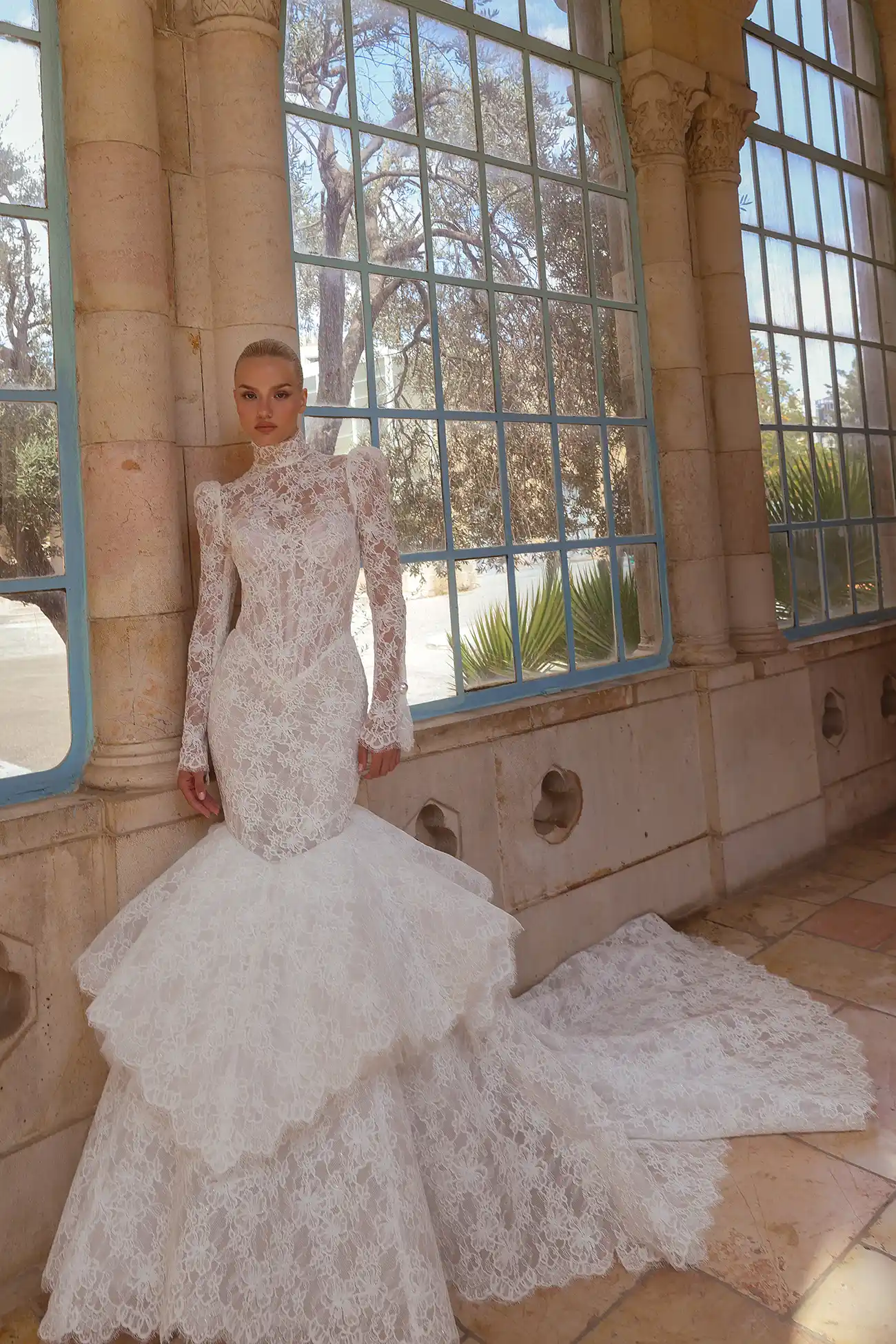 High-neck alençon lace mermaid gown with basque waist, tiered skirt, long puff sleeves, and pearl buttons.