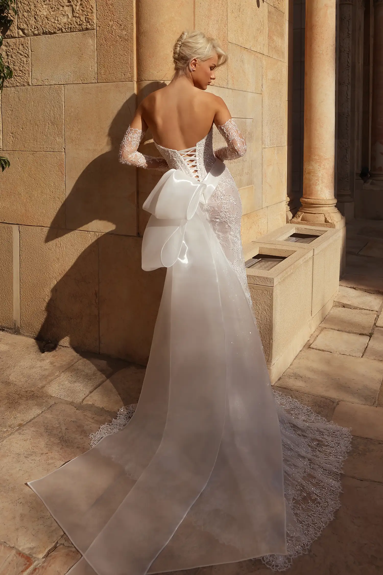 Hand-beaded chantilly lace sheath gown with high-shine liquid organza draping at neckline and basque waist and signature Pnina bow. Shown with beaded chantilly lace gauntlet gloves. 