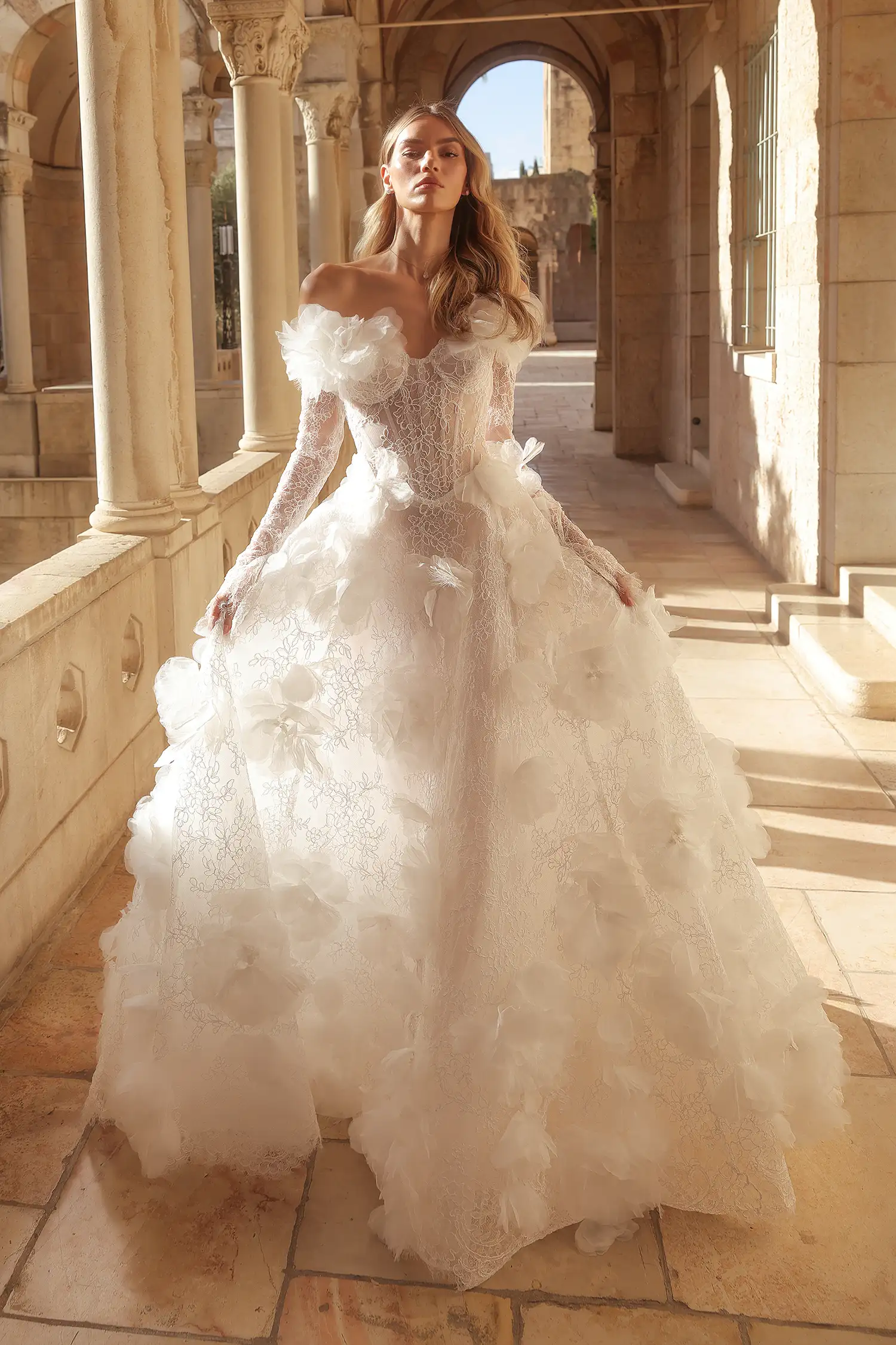 Alençon lace A-line gown with off-the-shoulder sweetheart neckline, long sleeves, and organza dimensional flowers with pearl and feather details.