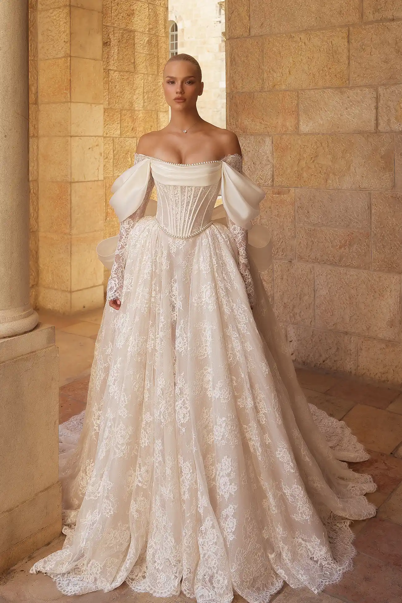 Alençon lace long-sleeved ballgown with silk organza draping at neckline and sleeves, exposed satin boning, pearl details at neckline and waistline, and signature Pnina silk organza bow. 