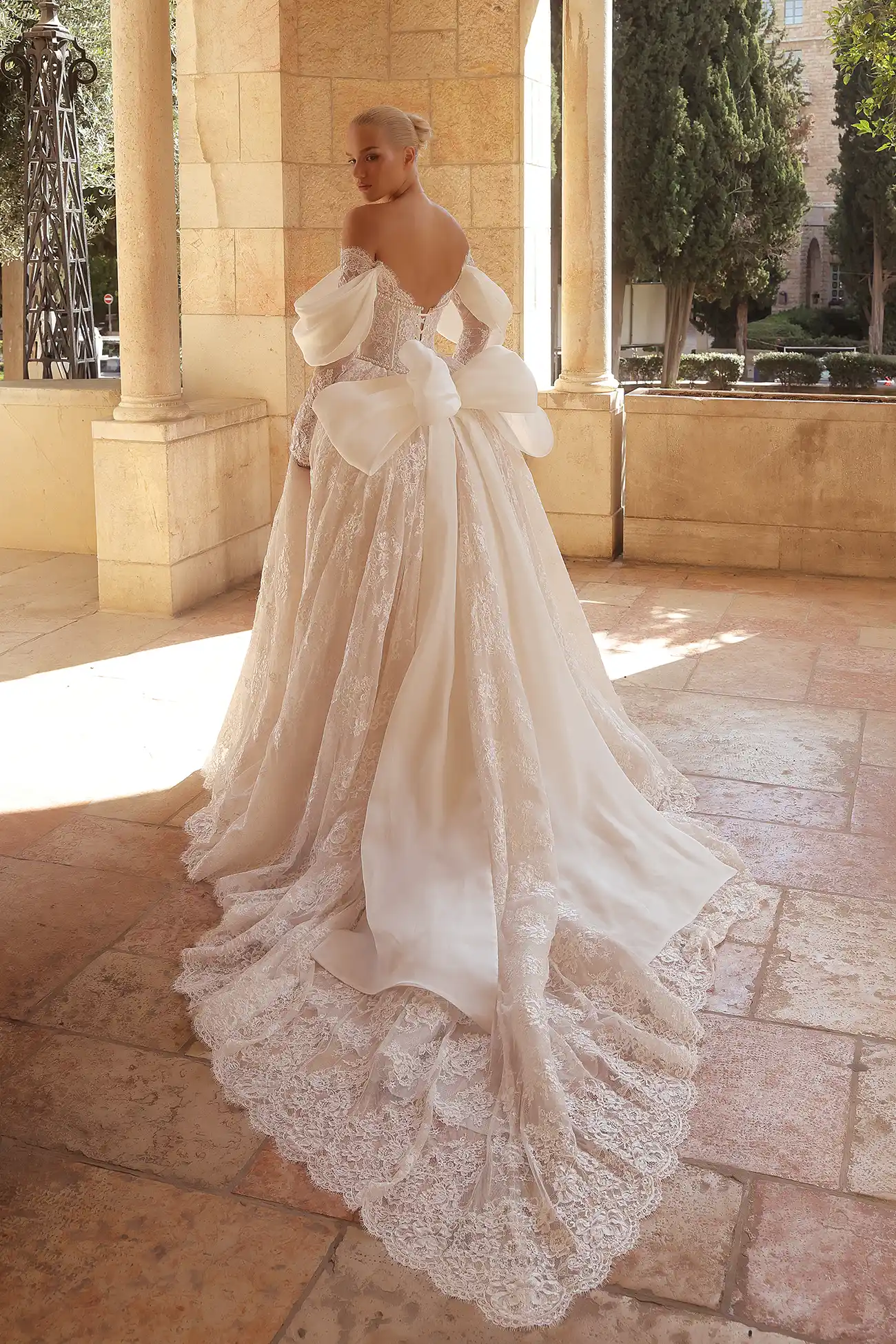 Alençon lace long-sleeved ballgown with silk organza draping at neckline and sleeves, exposed satin boning, pearl details at neckline and waistline, and signature Pnina silk organza bow. 